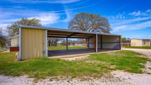A large metal carport with a corrugated roof stands on a concrete slab next to a gravel driveway, surrounded by green grass and trees under a bright blue sky.