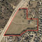 Aerial view of a large, open grassy area bordered by a red outline, surrounded by roads, trees, scattered buildings, and residential neighborhoods. The outlined area has a few trees and small structures within it.