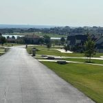 A paved road winds through the quiet Aledo neighborhood, 76008, with well-kept lawns, modern houses, and trees, overlooking a distant lake under a hazy sky.