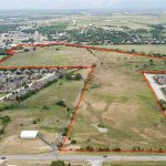 Aerial view of a large, undeveloped grassy plot of land outlined in red, surrounded by residential neighborhoods and roads, with more buildings and greenery visible in the distance.