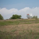 A grassy field with sparse trees and a white water tower in the background labeled "Aledo" under a partly cloudy sky.