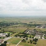 Aerial view of a suburban area with clusters of houses, roads, and buildings surrounded by open fields and greenery under a cloudy sky. The landscape extends into the horizon.