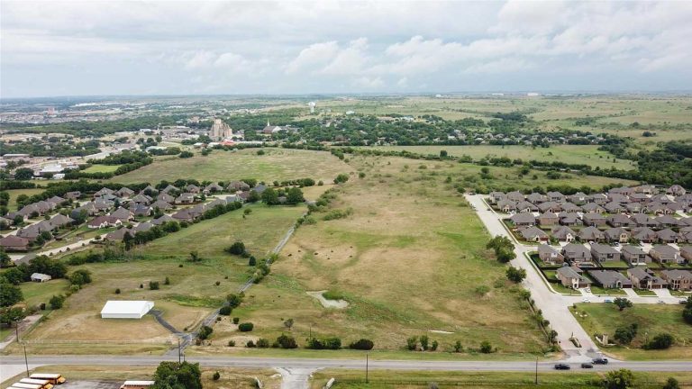 Aerial view of a suburban area with clusters of houses surrounding a large open grassy field, sparse trees, and roads under a partly cloudy sky.