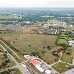 Aerial view of a large open field outlined in red, surrounded by residential neighborhoods, roads, and scattered buildings in a semi-rural area with green landscape and distant horizon.