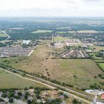 Aerial view of a suburban area with clusters of houses, roads, open green fields, and scattered trees under a cloudy sky, showing a mix of residential and undeveloped land.