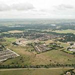 Aerial view of a suburban neighborhood with clusters of houses, patches of green fields, scattered trees, and a train traveling along tracks in the foreground under a cloudy sky.