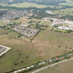 Aerial view of a suburban area with clusters of houses, open grassy fields, a school complex, and a long train moving along tracks near the bottom of the image. Trees and rolling hills extend into the distance.