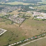 Aerial view of a suburban neighborhood with houses, schools, open fields, and a train traveling along tracks at the edge of the area, surrounded by greenery and trees.