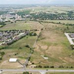 Aerial view of a suburban area with two neighborhoods of houses separated by an open, grassy field; roads and scattered buildings surround the residential zones, with more greenery and development in the distance.