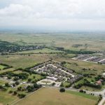 Aerial view of a suburban neighborhood surrounded by open fields and greenery under a cloudy sky, with roads and scattered buildings visible throughout the landscape.