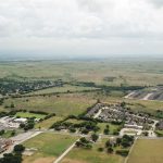 Aerial view of a suburban area with residential neighborhoods, scattered houses, roads, grassy open fields, and distant rolling hills under a cloudy sky.
