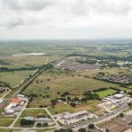 Aerial view of a suburban area with scattered houses, commercial buildings, roads, and large green fields extending into the distance under a cloudy sky. A railway or road runs diagonally across the scene.