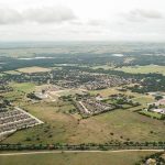 Aerial view of a suburban area with scattered houses, green fields, wooded areas, and a colorful train running along tracks in the foreground under a cloudy sky.