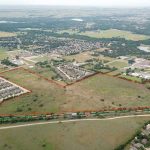 Aerial view of a suburban neighborhood with houses, open land, and trees. A large, irregularly shaped plot of land is outlined in red, separating it from surrounding residential areas and a nearby train track.