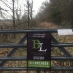 A metal gate blocks a narrow, grassy path surrounded by leafless trees. A real estate sign for Burt Ladner Real Estate, with contact info and website, is attached to the gate.