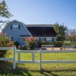 A blue barn-style house with a black roof sits behind a white wooden fence, surrounded by green grass, flowering bushes, trees, and a clear blue sky. Outdoor furniture is visible on a patio in front of the house.