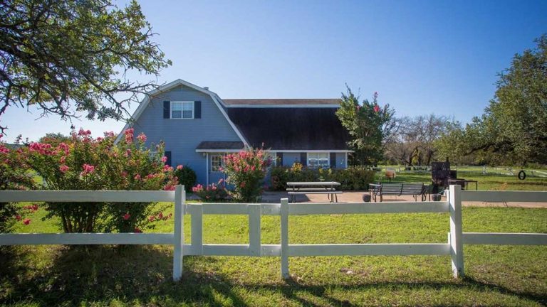A blue barn-style house with a black roof sits behind a white wooden fence, surrounded by green grass, flowering bushes, trees, and a clear blue sky. Outdoor furniture is visible on a patio in front of the house.
