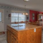 A spacious kitchen with wooden cabinets and a large island, featuring a built-in cooktop. There’s a door and two large windows, letting in natural light. The adjacent dining area has red walls and a wooden dining table with chairs.