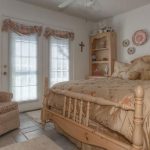 A cozy bedroom with a wooden bed, floral bedding, a beige armchair, and a shelf with decor. French doors with sheer curtains and a window bring in natural light. Pastel accents and wall decorations add charm.