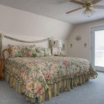 A bright bedroom with a floral-patterned bedspread, matching pillows, two bedside tables with lamps, framed wall art, a ceiling fan, and double glass doors letting in natural light.