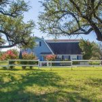 A gray barn-style house with a white fence sits behind blooming bushes and two large, leafy trees on a grassy lawn under a clear blue sky.