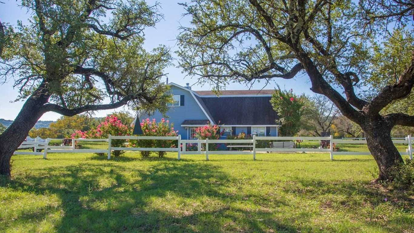 A gray barn-style house with a white fence sits behind blooming bushes and two large, leafy trees on a grassy lawn under a clear blue sky.