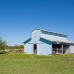 A light blue barn with a metal roof stands in a grassy field under a clear blue sky, with trees and farm equipment visible in the background.