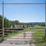 A rustic metal gate with "ROCK N CEDAR" written on it stands at the entrance to a green, open field with trees and blue sky in the background.