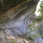 A large, curved rock overhang extends above a shaded, rocky area with green foliage and tree branches surrounding the scene. Sunlight filters through the leaves, creating a mix of light and shadow.