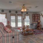 Living room with patterned sofas, a coffee table, and a large TV. Decor includes deer mounts, a "TEXAN" sign, western star accents, and a telescope near large windows with floral curtains overlooking greenery.