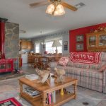 Cozy living room with plaid sofa, armchair, wooden coffee table, and red stove by a stone wall. Dining table is visible near kitchen. Decor includes floral accents and a red accent wall with cabinets. Ceiling fan above.