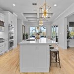 Spacious modern kitchen with white cabinets, a large island, pendant lights, stainless steel appliances, light wood flooring, and large windows letting in natural light.