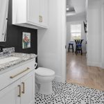 Modern bathroom with white cabinets, gold handles, granite countertop, and a white toilet. The floor has black and white hexagonal tiles. A hallway with light wood flooring leads to a dining area with a table and chairs.