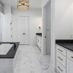 Modern bathroom with black and white patterned wall, marble flooring, large bathtub with black fixtures, white cabinets with gold handles, a large mirror, and gold pendant lights.