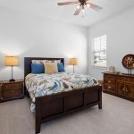 A bedroom with a wooden bed frame, colorful bedding, two bedside tables with lamps, a ceiling fan, a large wooden dresser with decor, and a window with white shutters. The walls are white and the carpet is beige.