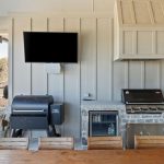 Outdoor kitchen with a wood dining table, grill, smoker, mini fridge, sink, and a wall-mounted TV. Stonework surrounds the grill area, and the space opens to a yard with trees visible in the background.
