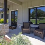 A front porch with two dark wicker chairs and a small table, bordered by stone walls and pillars, overlooks a green lawn and trees, with sunlight casting shadows across the area.