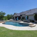 Backyard view of a modern house with a metal roof, pool, attached hot tub, outdoor seating, green lawn, and trees under a clear blue sky.