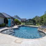A backyard with an in-ground swimming pool, hot tub, lounge chairs, patio umbrella, and a glass greenhouse under a clear blue sky. Trees and houses are visible in the background.