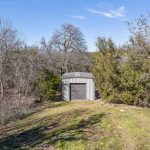 A small gray metal shed with a closed roll-up door sits on a grassy clearing surrounded by leafless and evergreen trees under a blue sky.