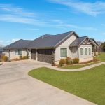 Single-story house with gray siding, dark roof, and large driveway, surrounded by green lawn and open landscape under a bright blue sky with scattered clouds.