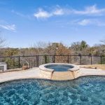 A backyard swimming pool with a connected circular hot tub, surrounded by a metal fence and overlooking trees under a blue sky with wispy clouds.