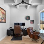 A modern home office with a brown leather chair at a glass desk, dual monitors on a black desk, abstract art on white walls, and large window letting in natural light.