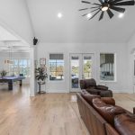 Spacious open-concept living area with brown leather sofas, stone fireplace, mounted TV, large windows, kitchen with bar stools, dining area, pool table, and view of a backyard patio and trees.