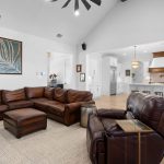 Spacious living room with brown leather sectional sofa and armchair, neutral rug, wall art, and an open layout leading to a modern white kitchen with island seating. Natural light brightens the area.