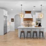 Modern kitchen with white cabinets, stainless steel appliances, a central island with three stools, pendant lights, and a wood-accented range hood. A pool table is visible in the adjoining room.