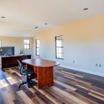 Spacious office with wood flooring, beige walls, large windows, and a wooden desk with a computer and chair. The room is mostly empty with plentiful natural light.
