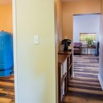 A hallway with wood-patterned flooring leads to a living room with a couch and TV. On the left, an open door reveals a bathroom with a toilet and a large blue tank. A bookshelf and coffee maker are in the hallway.