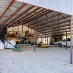 A large open metal shed with a gravel floor houses a white pickup truck, a golf cart, stacked shelves with tools and supplies, large white storage bags, and various equipment. Trees and grass are visible outside.