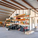 A spacious metal garage with exposed beams, concrete floor, and large shelves storing tools, equipment, and containers. A workbench, ladders, boxes, and various items are arranged neatly throughout the space.